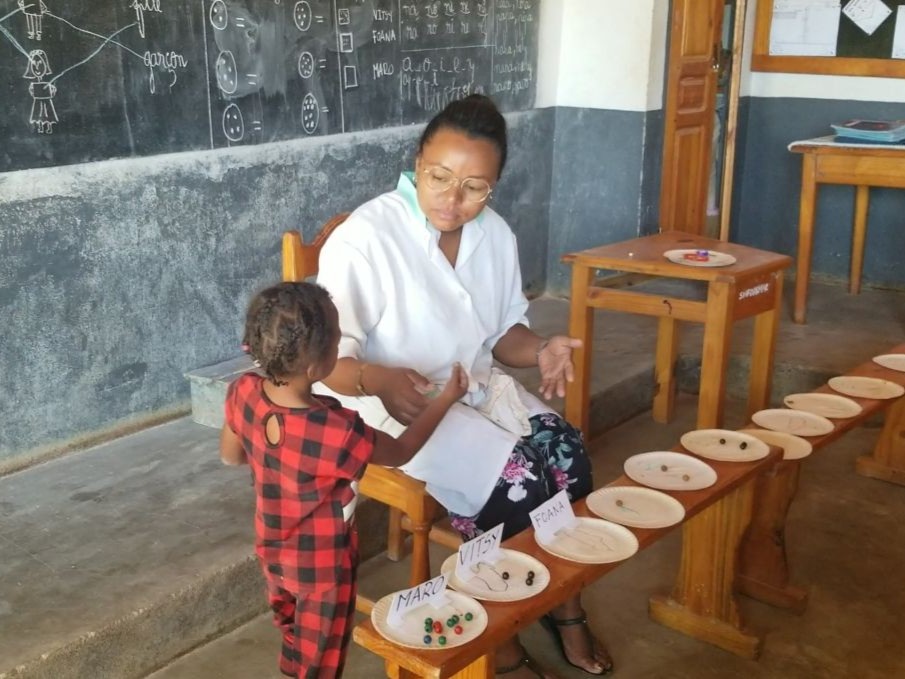 A female teacher sits behind paper plates with various quantities of marbles. She encourages a 5-year-old student to use sign language to describe the quantities of items displayed.
