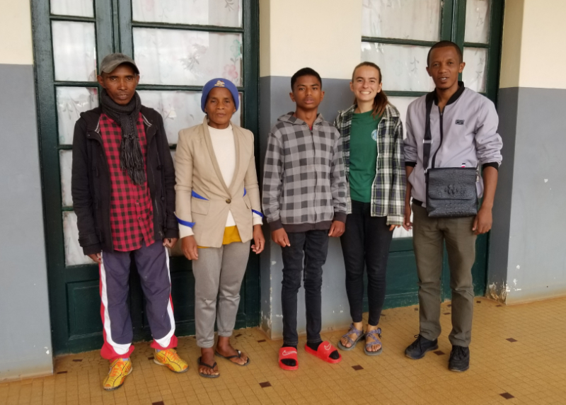 Five people pose for a photo outside a concrete hospital building. Mandimby's father and mother stand to the right. His father wears a baseball cap, a black scarf, and a open zippered jacket. His mother wears a winter hat with a professional-looking tan business jacket. Mandimby, center, is a thin teenage boy slightly taller than 5 feet in height. He wears a checkered zip-up and looks healthy. His face is unsmiling due to partial facial paralysis, the result of an illeness when he was an infant. Vanessa is next, smiling brightly in her chacos and L.L. Bean flannel. Finally, the school director wears a grey zip-up with dark, professional pants and a over-the-shoulder bag which carries paperwork and other important materials.