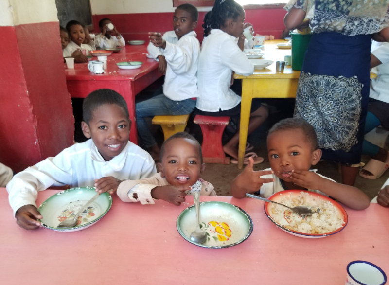 Three boys sit next to each other at a pink wooden tables. Two of the plates are empty, while the third still has rice and beans in it.