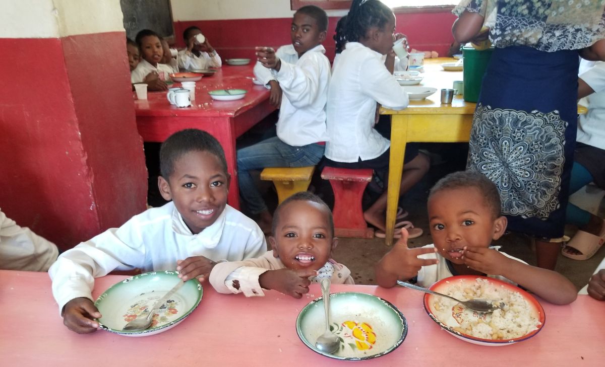 three young children smile for a photo while sitting at a dining table in a cafeteria