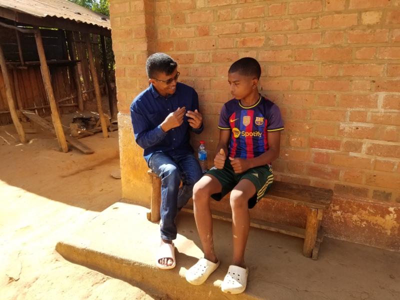 SEMAFI's cook, Liva, sits on a wooden bench with Mandimby outside his home. Liva's hands form the Malagasy sign for "vocational training" as he discusses the program with Mandimby.