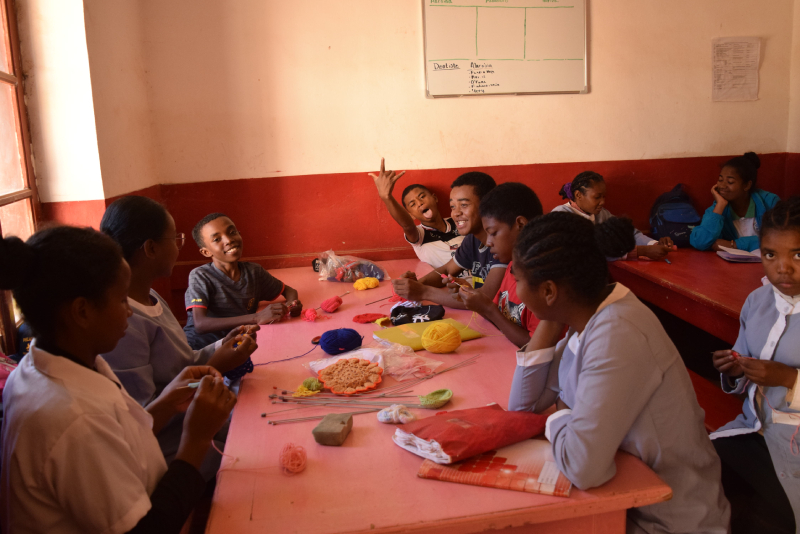 Eight vocational training students and their teacher practe crochet and knitting in SEMAFI's dining room. At the end of the table, three teenage boys appear to be slacking. One of them is Mandimby, who sticks his tounge out and has his hand up, his thumb, pointer finger, and pinky extended for the camera.