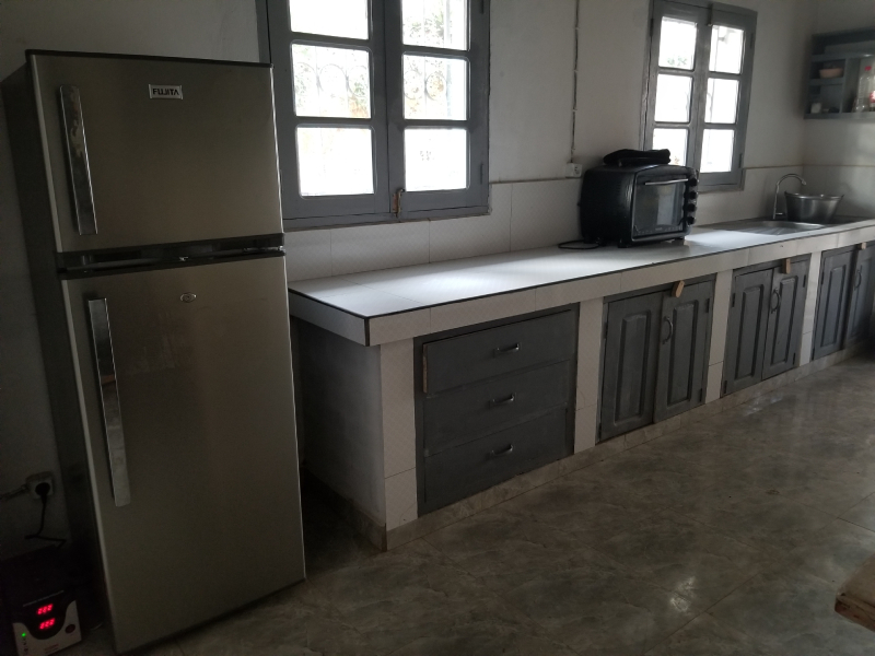 A photo of the new kitchen. To the left is a electricity stabilizer and a medium size refridgerator. To the right of the refridgerator is a long kitchen counter overlayed with tiles. There are cabinets for storage underneath the counter. On the counter is a small toaster oven. At the end of the counter is a small sink. On the wall is two openable windows with glass panes. The floor is tile.